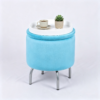 Light blue velvet round storage ottoman with metal legs, featuring a white wooden tray top holding a coffee cup and succulent plant on a plain background.