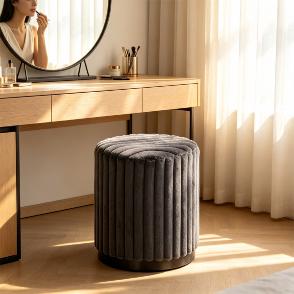 Dark gray pleated velvet round storage ottoman stool styled in front of a wooden vanity desk in a sunlit room.