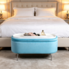Light blue velvet oval ottoman storage bench placed near a bed with warm beige bedding and cozy neutral decor.
