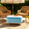 Light blue velvet oval storage ottoman set up on a sunlit patio with wooden outdoor lounge chairs.